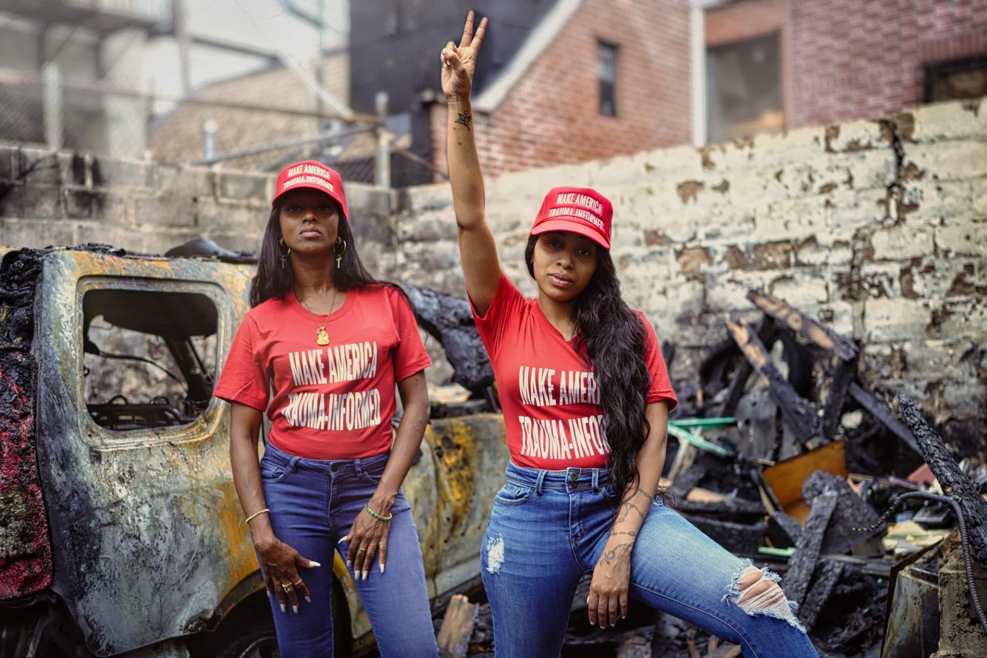 Two black women pose confidently in the middle of a pile rubble. They wear red t-shirts and baseball caps reading "Make America Trauma-Informed". One woman holds her arm above her head making the peace symbol with her hand.